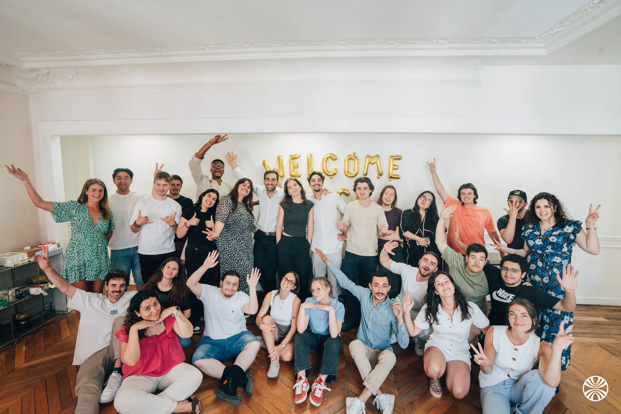 Groupe de jeunes adultes souriants posant ensemble dans une pièce avec des ballons dorés formant le mot « WELCOME » au mur.