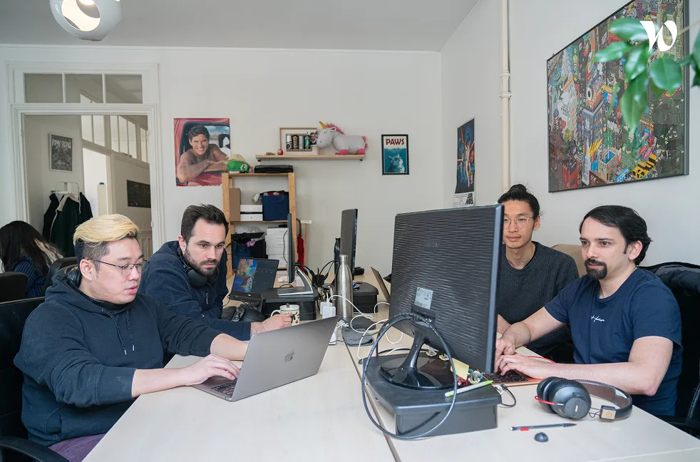 Quatre personnes concentrées travaillant sur des ordinateurs à une grande table dans un bureau moderne avec des décorations murales colorées.
