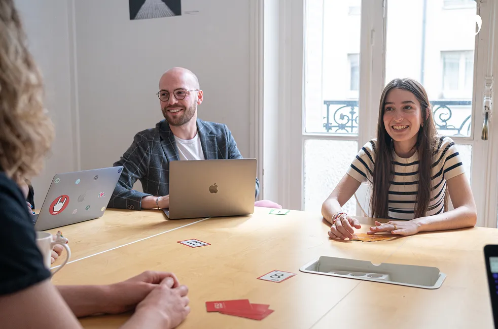 Quatre personnes souriantes autour d'une table en bois avec des ordinateurs portables et des cartes de jeu, dans une salle lumineuse avec une grande fenêtre.