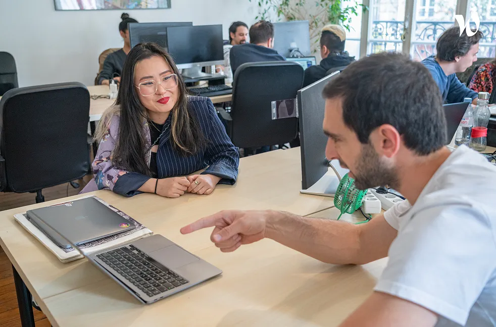 Un homme montre quelque chose sur un ordinateur portable à une femme souriante dans un bureau avec plusieurs collègues travaillant en arrière-plan.
