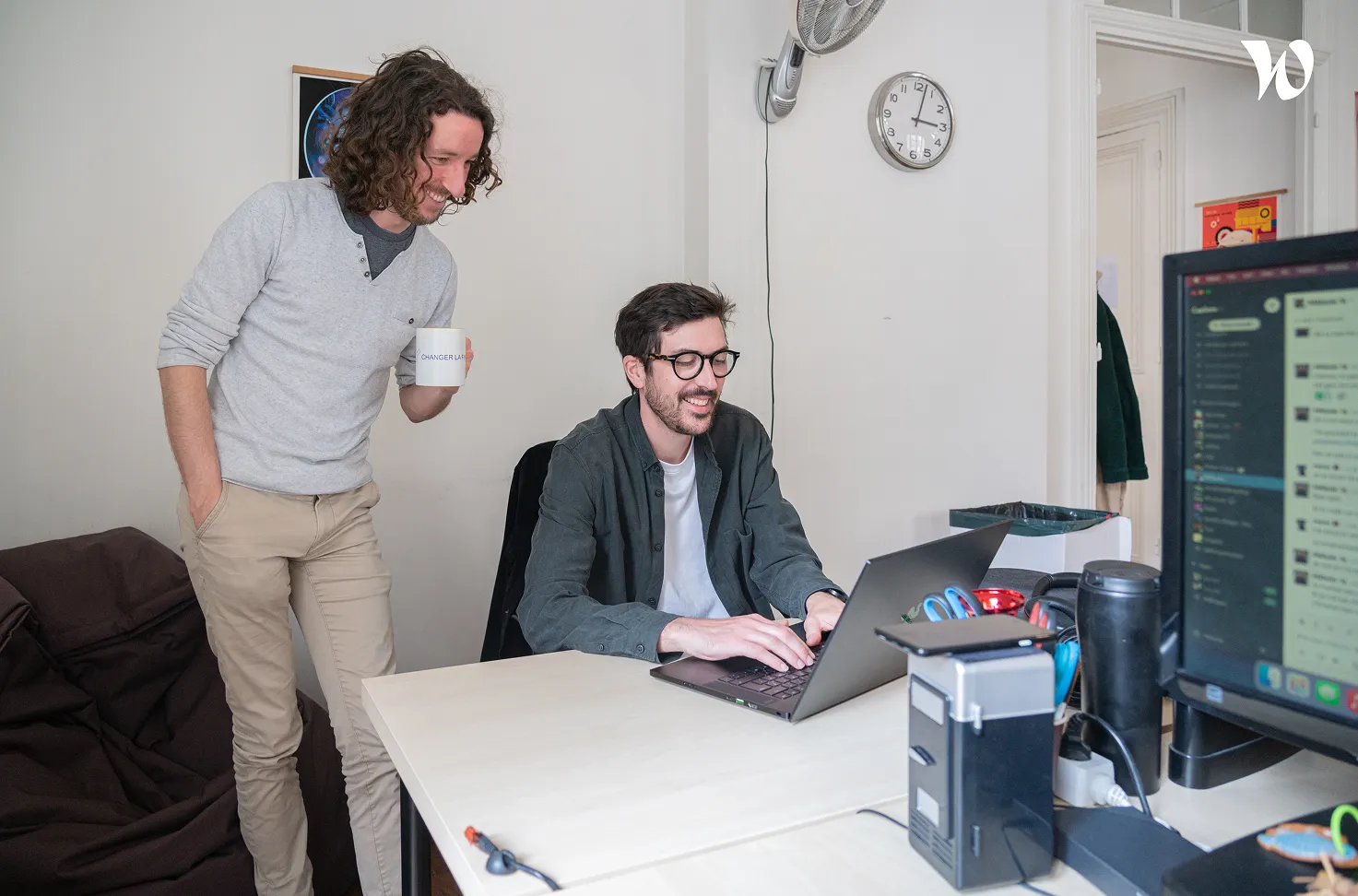 Deux hommes dans un bureau, l'un tape sur un ordinateur portable tandis que l'autre, tenant une tasse, le regarde en souriant.