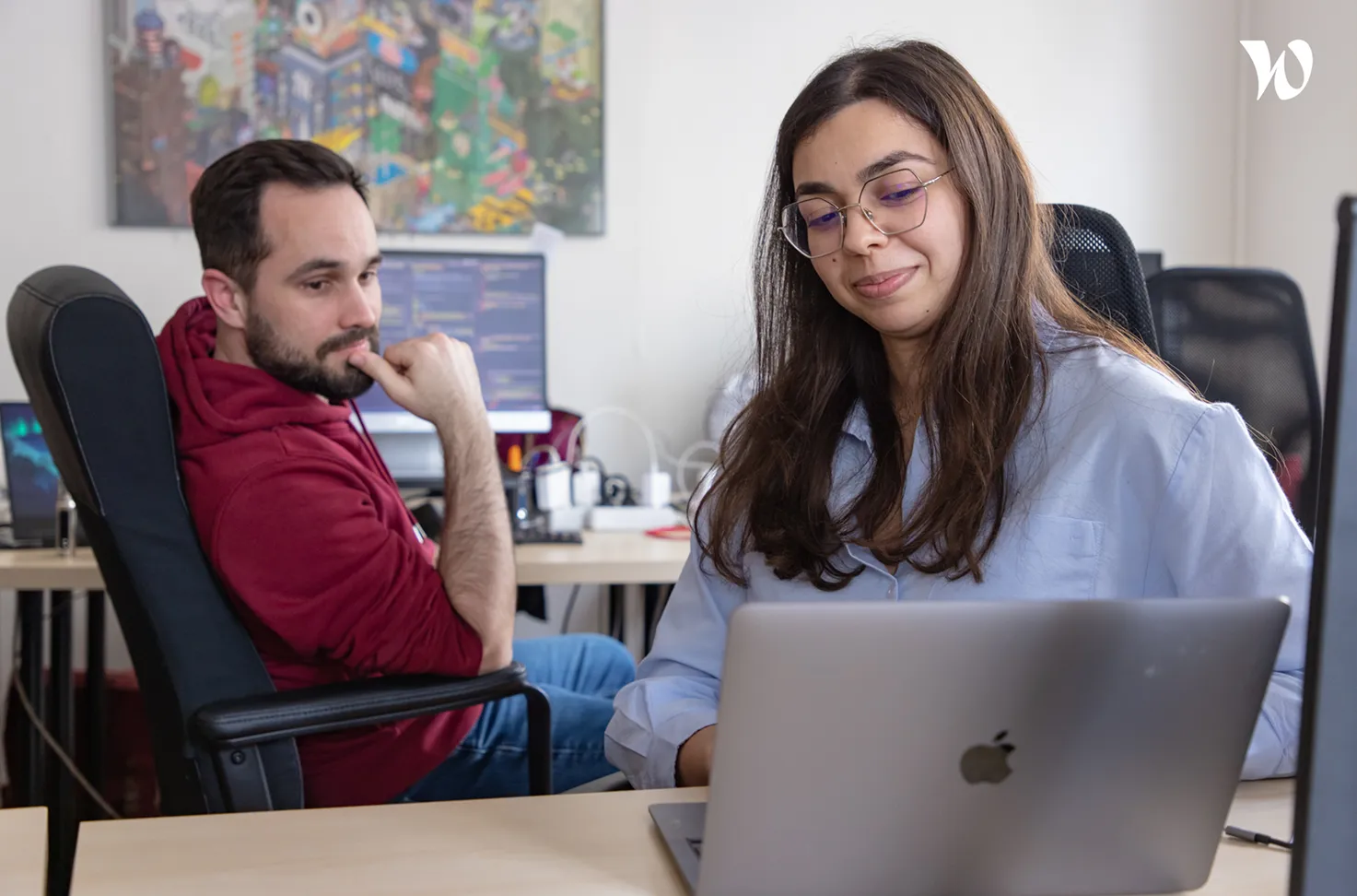 Deux collègues travaillant ensemble dans un bureau moderne, une femme souriante regarde un ordinateur portable Apple tandis qu'un homme en sweat à capuche rouge réfléchit en arrière-plan.