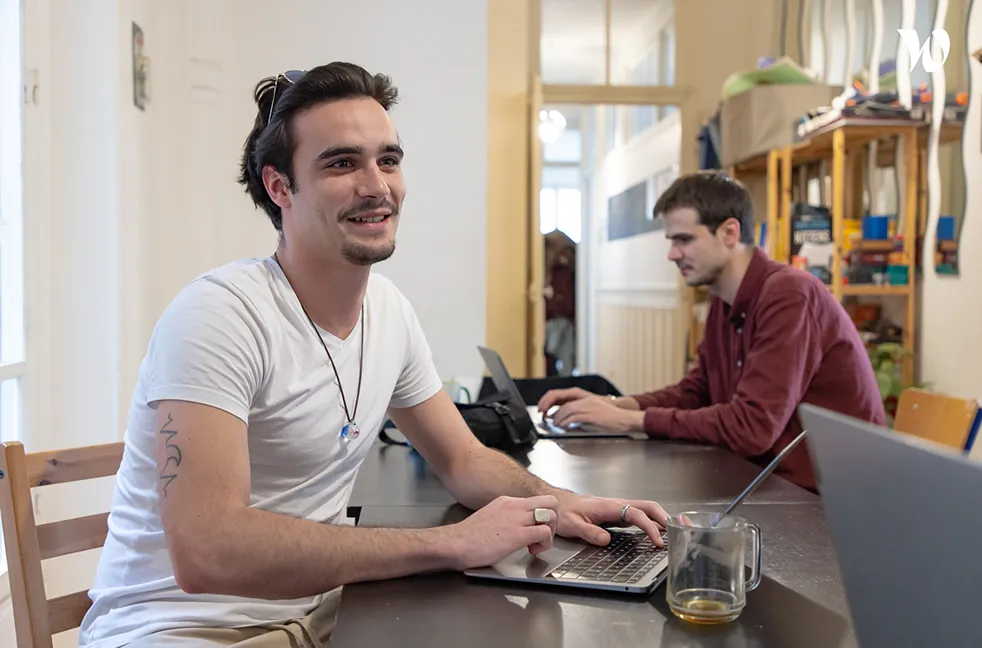 Jeune homme souriant, assis à une table avec un ordinateur portable, avec une autre personne travaillant sur un ordinateur en arrière-plan.