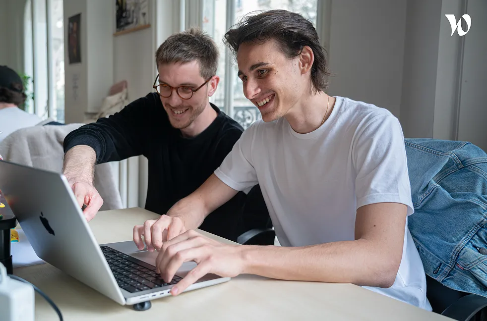 Deux jeunes hommes souriant travaillant ensemble sur un ordinateur portable dans un bureau lumineux.