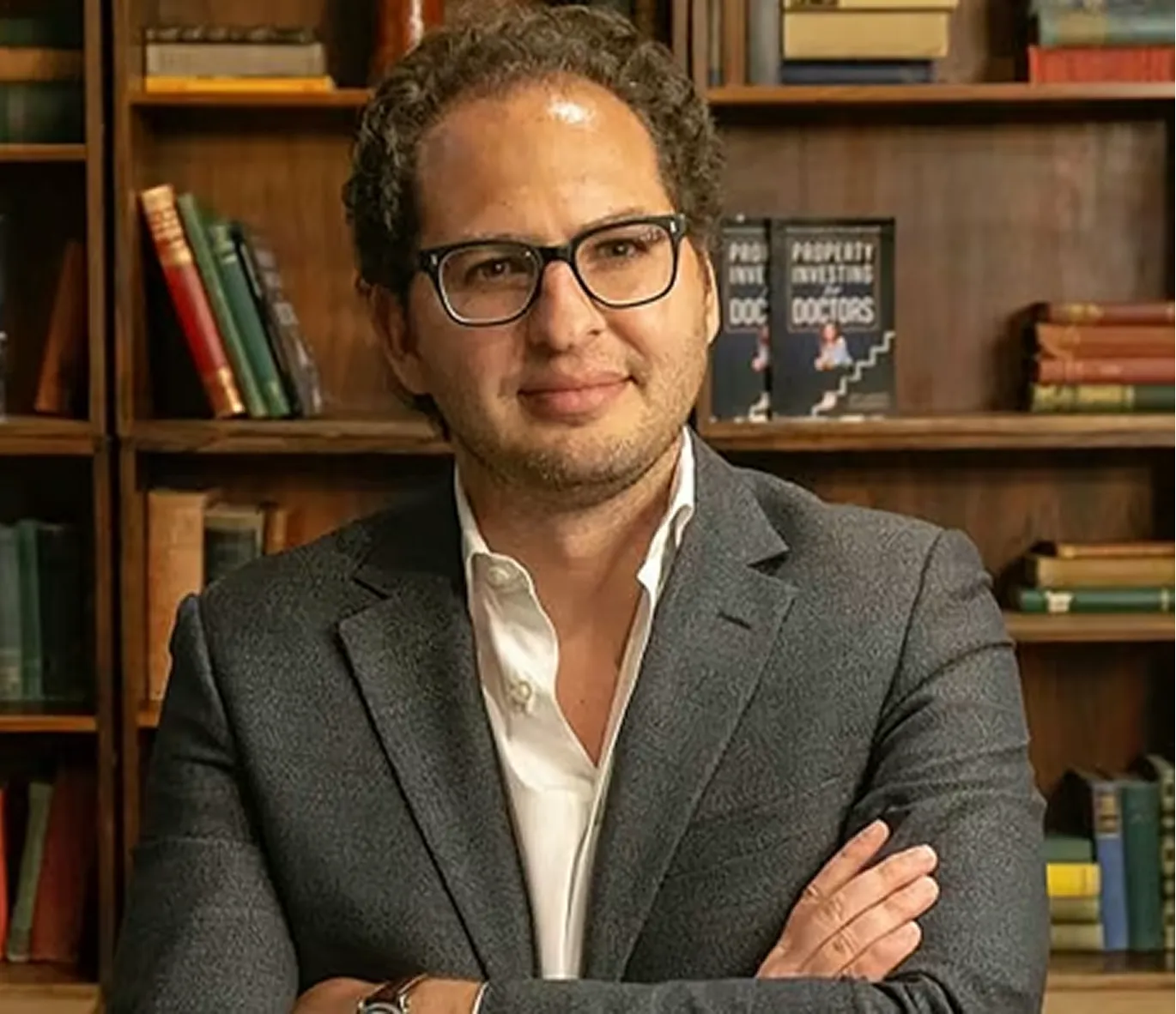 Homme portant des lunettes et un costume gris avec les bras croisés devant une bibliothèque remplie de livres.