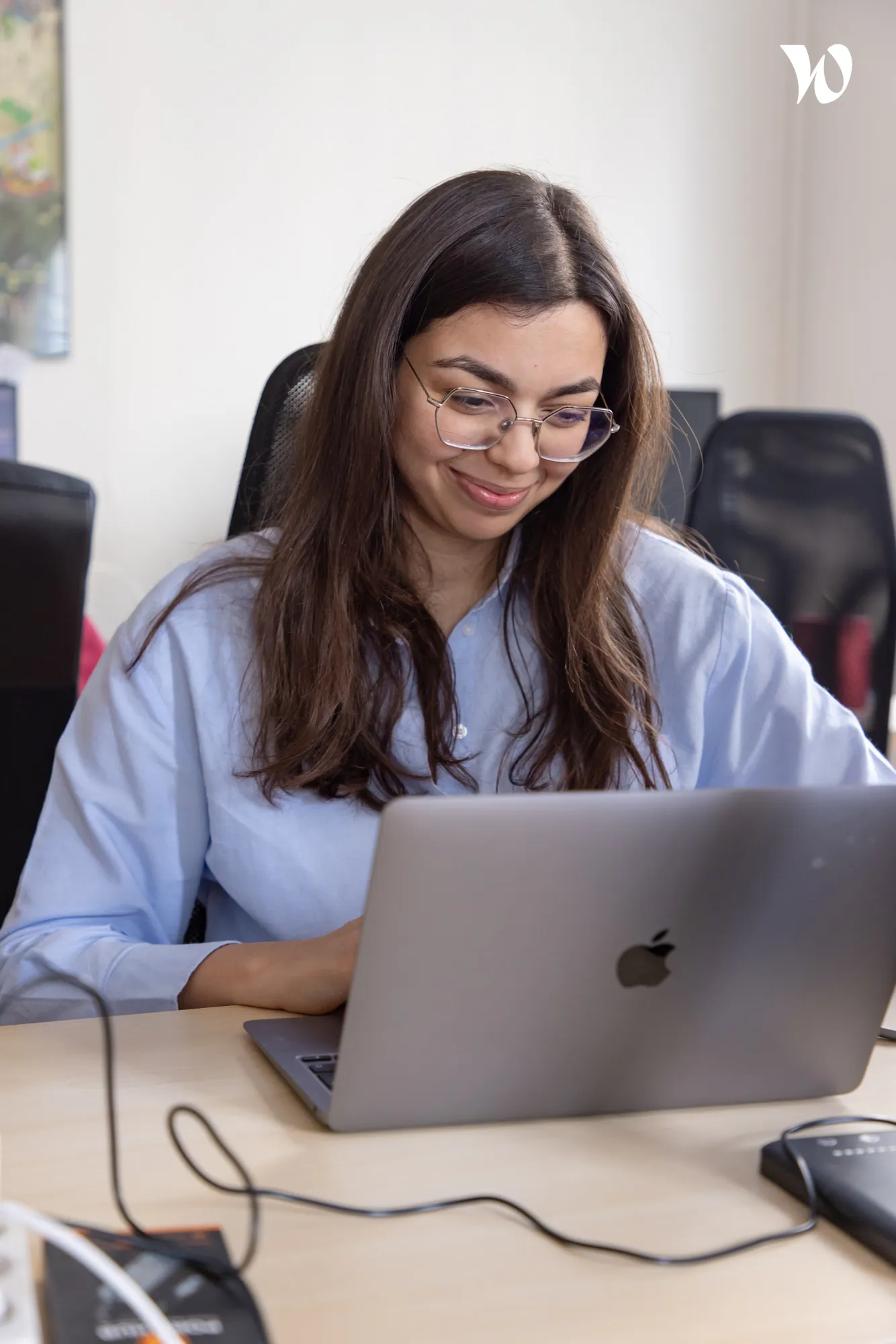Jeune femme souriante portant des lunettes et une chemise bleu clair, travaillant sur un ordinateur portable Apple dans un bureau moderne.