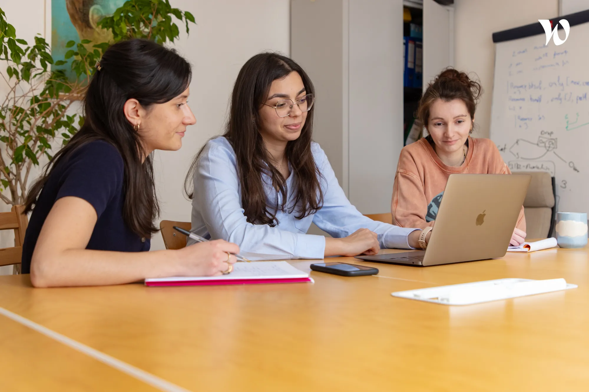 Trois femmes discutant et travaillant ensemble autour d'une table avec un ordinateur portable, un carnet et une tasse dans une salle de réunion.