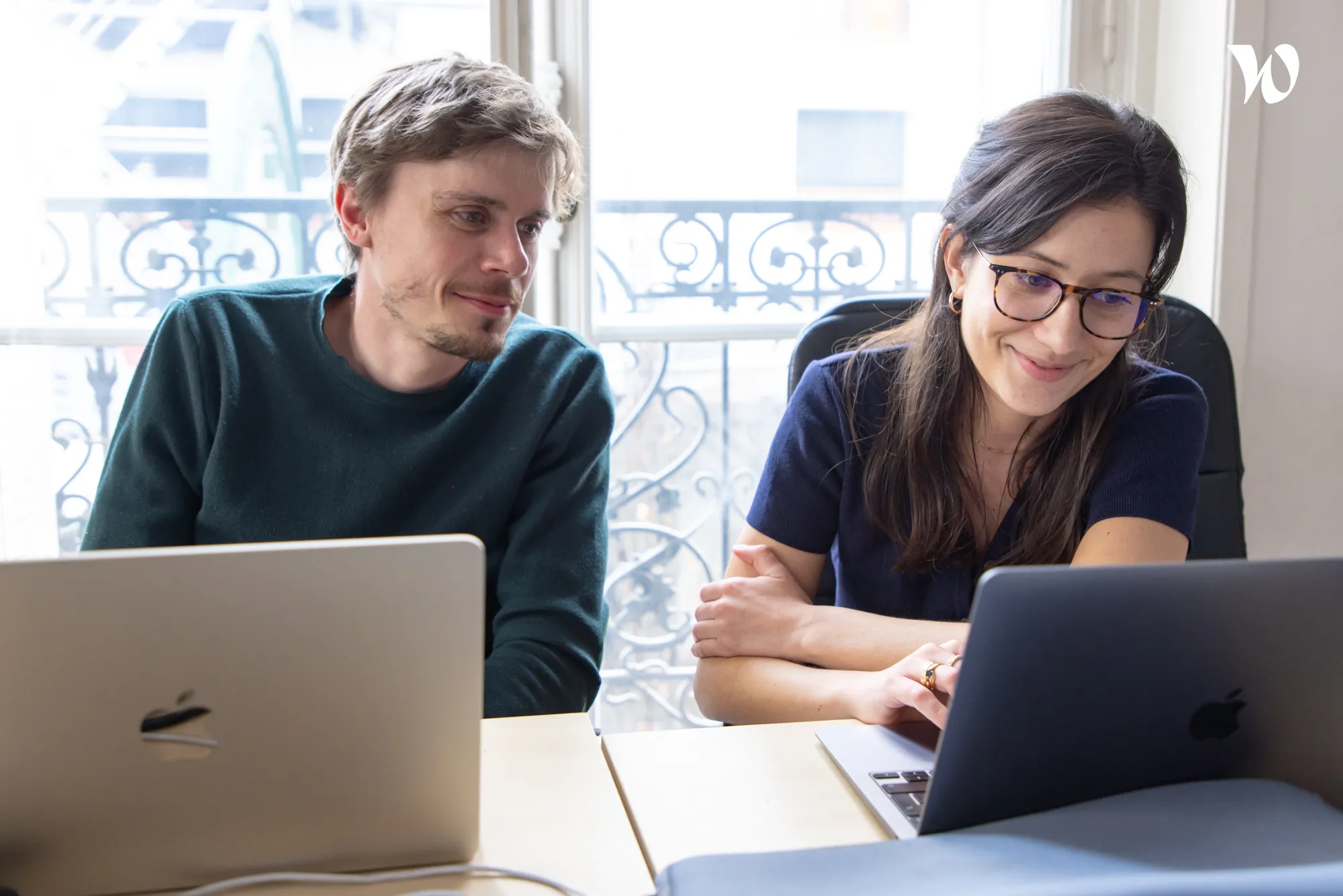 Un homme et une femme travaillent côte à côte avec des ordinateurs portables Apple dans un bureau lumineux avec une fenêtre ornée.