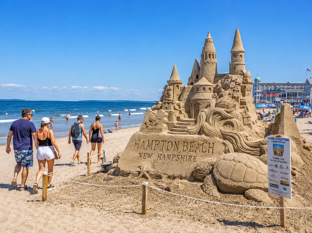A sunny summer scene at Hampton Beach, New Hampshire, showing detailed sand sculptures, beach visitors walking nearby, and the Atlantic Ocean in the background.
