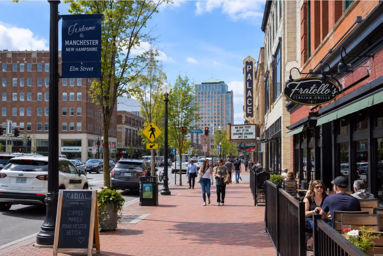 A street-level view of downtown Manchester, New Hampshire, along Elm Street with local storefronts, sidewalks, and city activity, representing the active Manchester–Nashua housing market and Southern NH lifestyle.