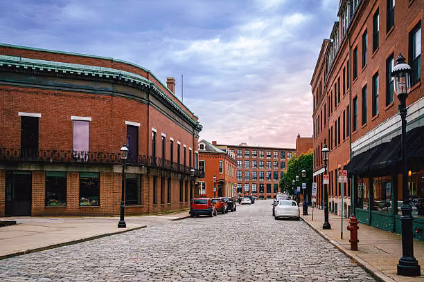 A street-level view of downtown Lowell, Massachusetts, showing historic mill buildings, sidewalks, and nearby canal scenery to reflect the city’s local character and walkable urban feel.