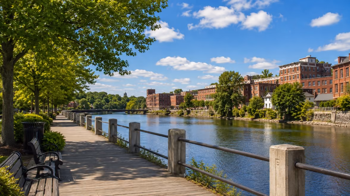 A realistic summer view of Haverhill, MA’s riverfront boardwalk near downtown, showing the Merrimack River, clean walking paths, benches, trees, and a welcoming New England community feel.