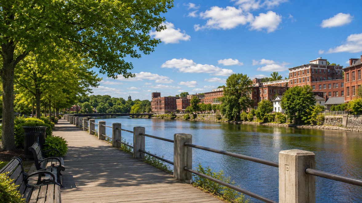 A realistic summer view of Haverhill, MA’s riverfront boardwalk near downtown, showing the Merrimack River, clean walking paths, benches, trees, and a welcoming New England community feel.