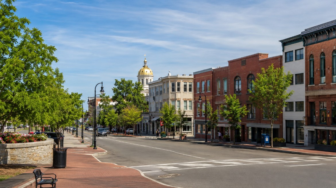 A realistic summer view of downtown Concord, NH, showing a clean walkable Main Street, trees, local storefronts, and a welcoming New England community feel.