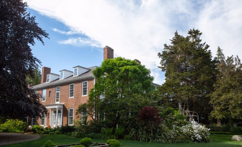 A realistic summer view of a North Andover, MA garden or historic community setting with mature trees, walking paths, and a calm New England residential feel.