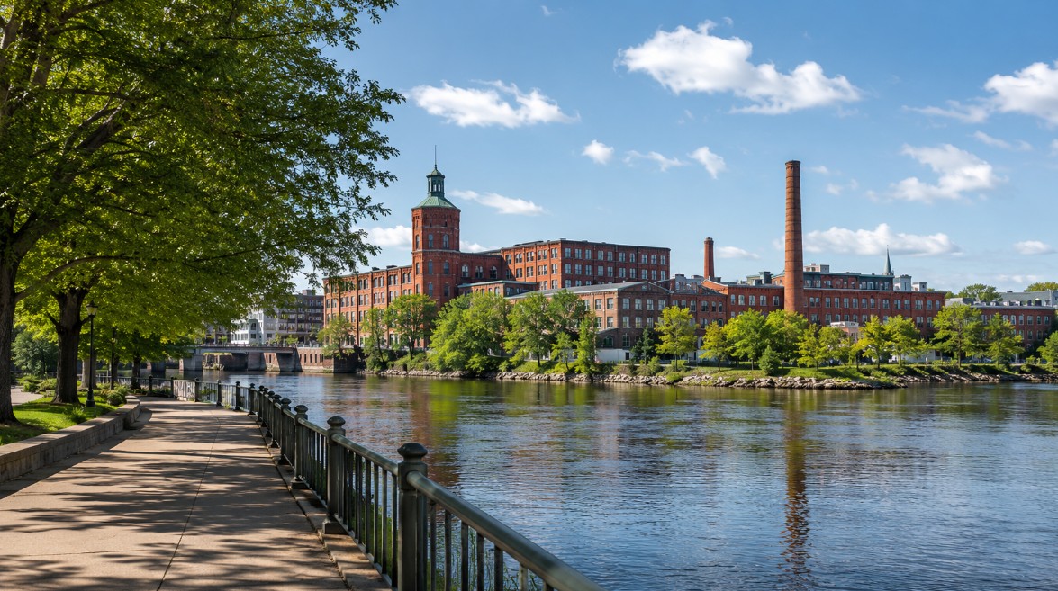 A realistic view of Lawrence, Massachusetts along the Merrimack River, showing historic red-brick mill buildings, mature trees, walkable riverfront paths, and a clean New England community feel.