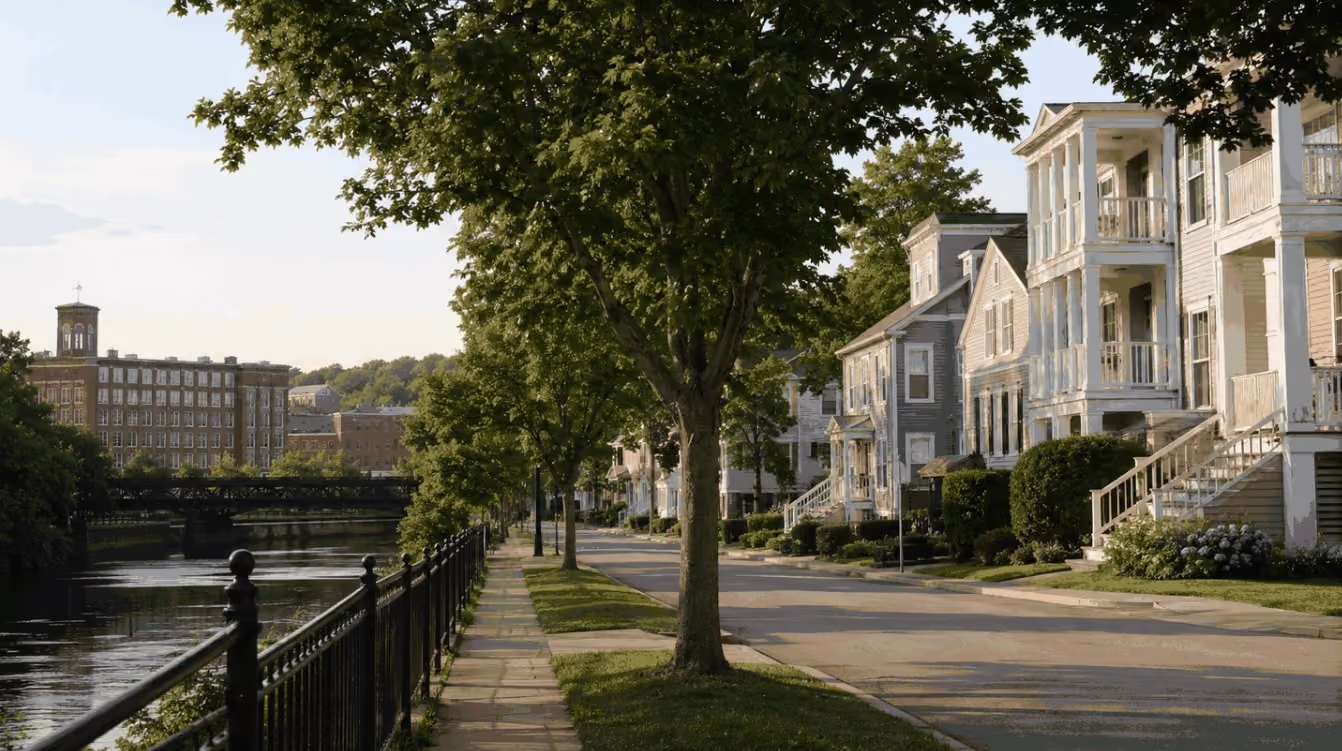 A realistic aerial view of an established Lowell, Massachusetts neighborhood with traditional New England homes, mature trees, residential streets, and the Merrimack River visible in the distance.