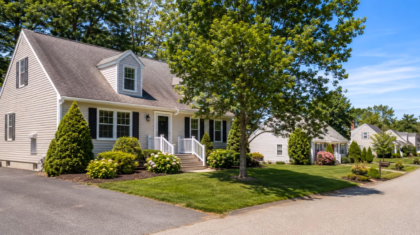 A polished but realistic Dracut, Massachusetts home in summer, with nearby New England-style homes subtly visible along the same residential street, illustrating how sellers compete for buyer attention through curb appeal and presentation