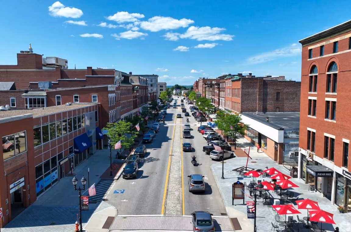 A sunny summer view of downtown Concord, New Hampshire, showing the State House’s gold dome, historic brick buildings, mature green trees, wide sidewalks, and a welcoming public green space.