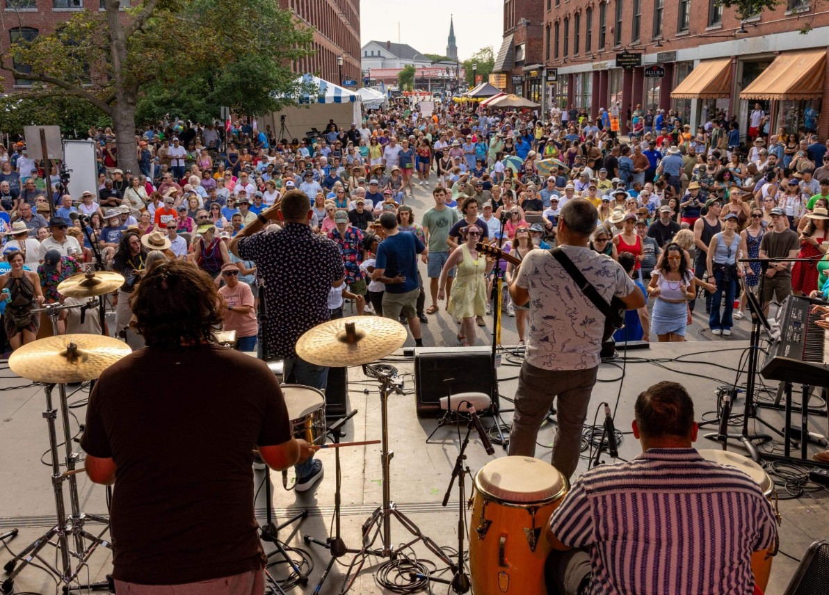 A lively summer folk festival scene in downtown Lowell, Massachusetts, with an outdoor performance, historic red-brick mill architecture, shaded seating, food tents, and a diverse crowd shown from a distance.
