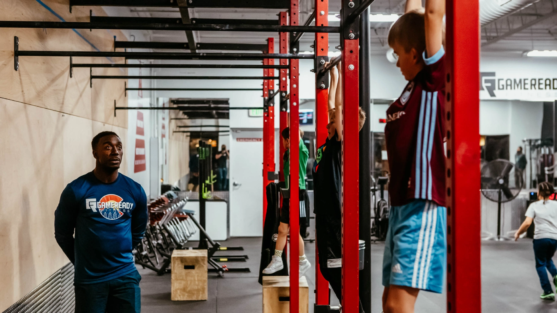 A man hangs from a pull-up bar in a gym at Game Ready Fitness in Thornton, CO, demonstrating strength training.