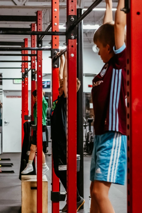 A man hangs from a pull-up bar in a gym at Game Ready Fitness in Thornton, CO, demonstrating strength training.