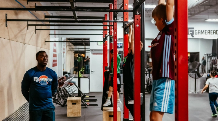 A man hangs from a pull-up bar in a gym at Game Ready Fitness in Thornton, CO, demonstrating strength training.