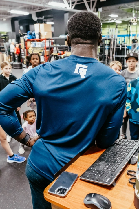 A man stands in front of a group of children in a gym at Game Ready Fitness in Thornton, CO, engaging with them.