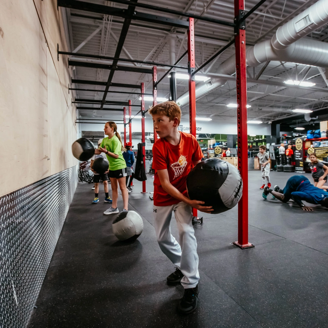 A young boy holds a large ball in a gym at Game Ready Fitness in Thornton, CO, smiling and ready to play.