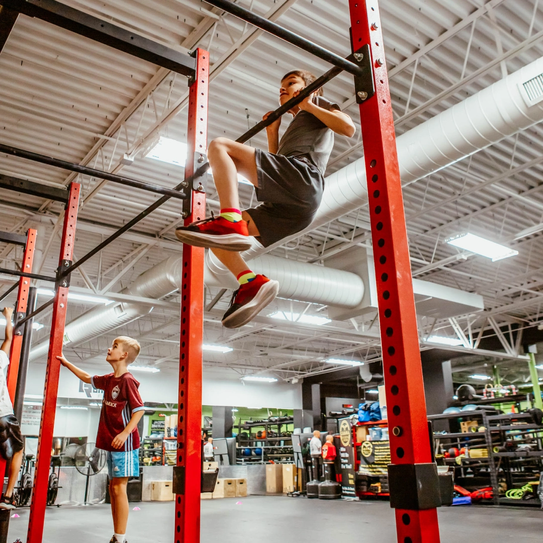 A boy performs a pull-up on a bar at Game Ready Fitness in Thornton, CO, showcasing strength and determination.