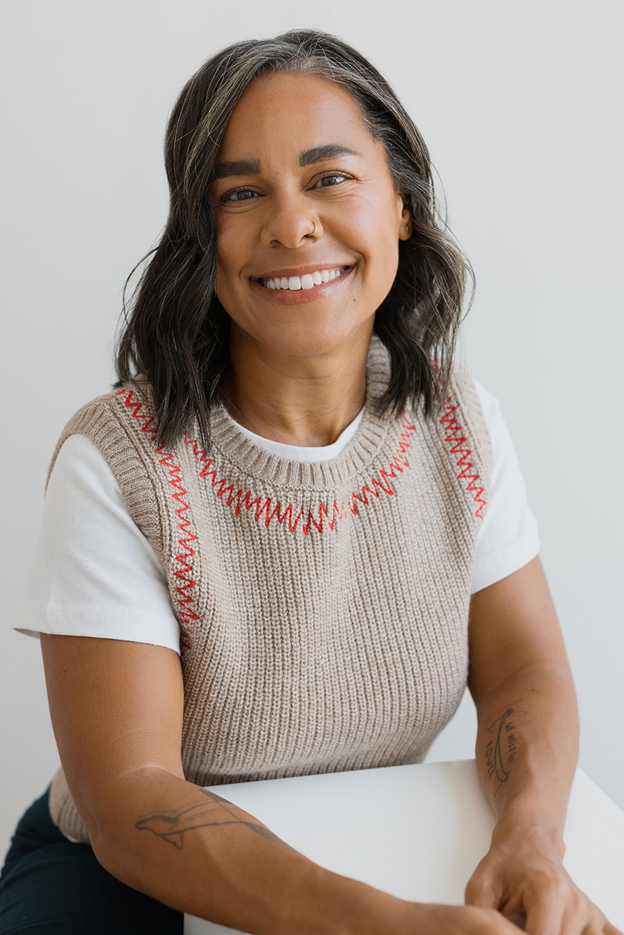 Smiling woman with shoulder-length dark hair wearing a beige knit vest with red stitching over a white t-shirt, sitting with arms resting on a white surface.