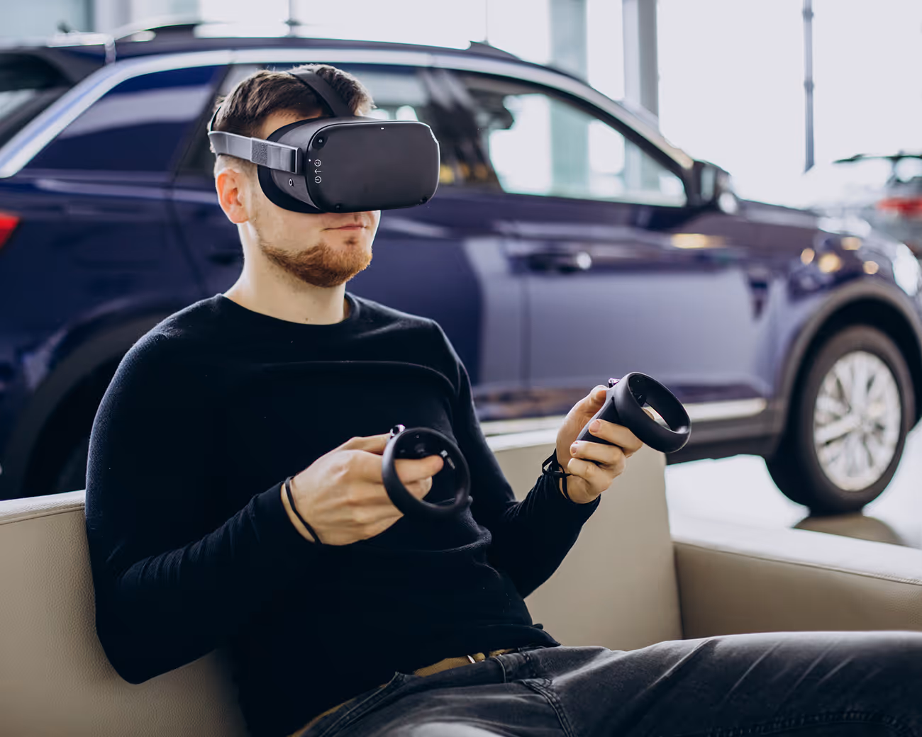 A man in a wearing a VR headset holding two controllers is sat in a car showroom with a car in the background