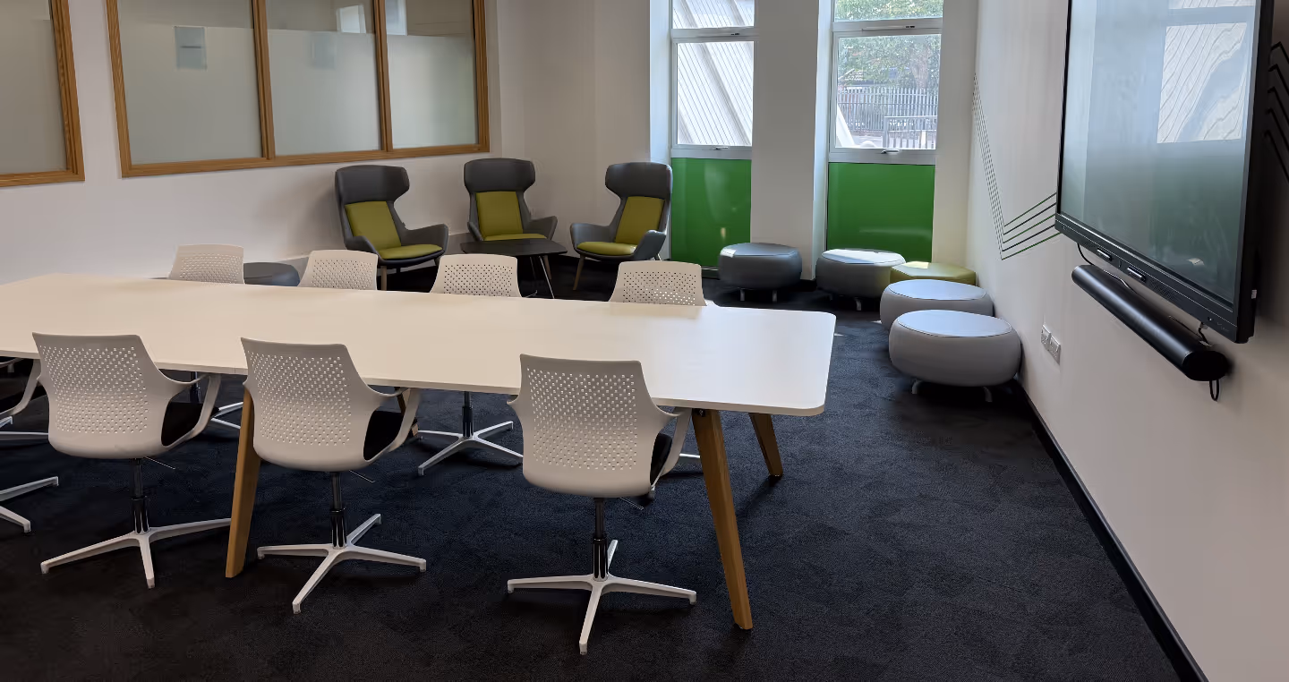 A room with a white table and 6 white chairs around. There is a tv screen on the wall and stools and other chairs in the background.