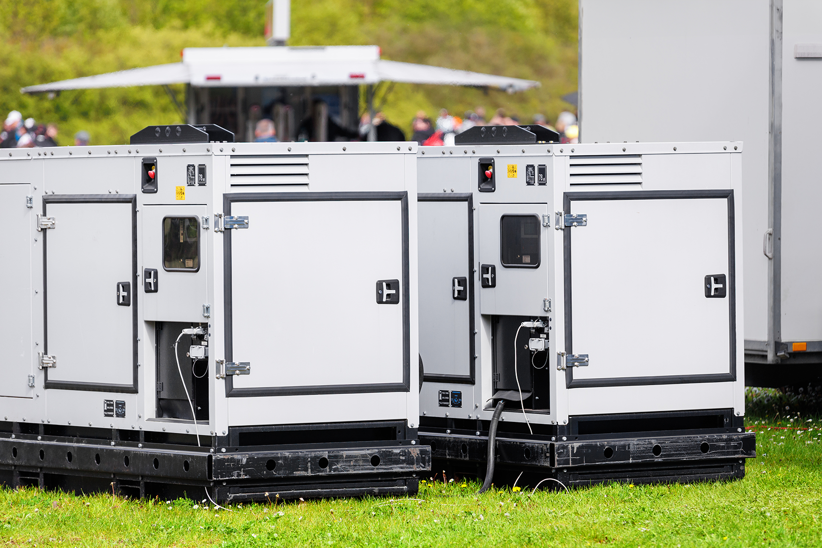 Two large white portable industrial power generators placed on grass with a blurred trailer and people in the background.