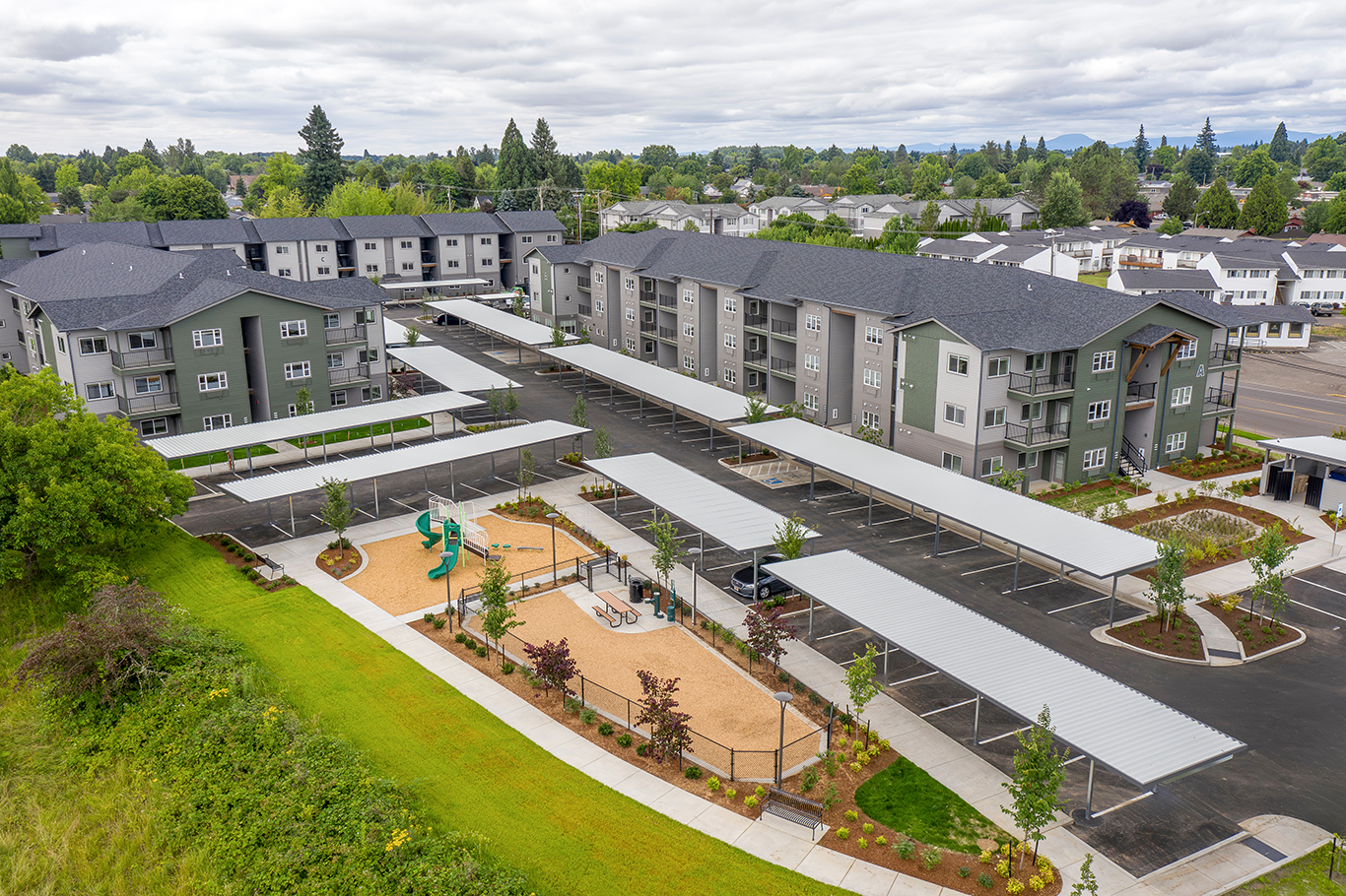 Aerial view of a modern apartment complex with covered parking and a fenced playground area with slides and seating.