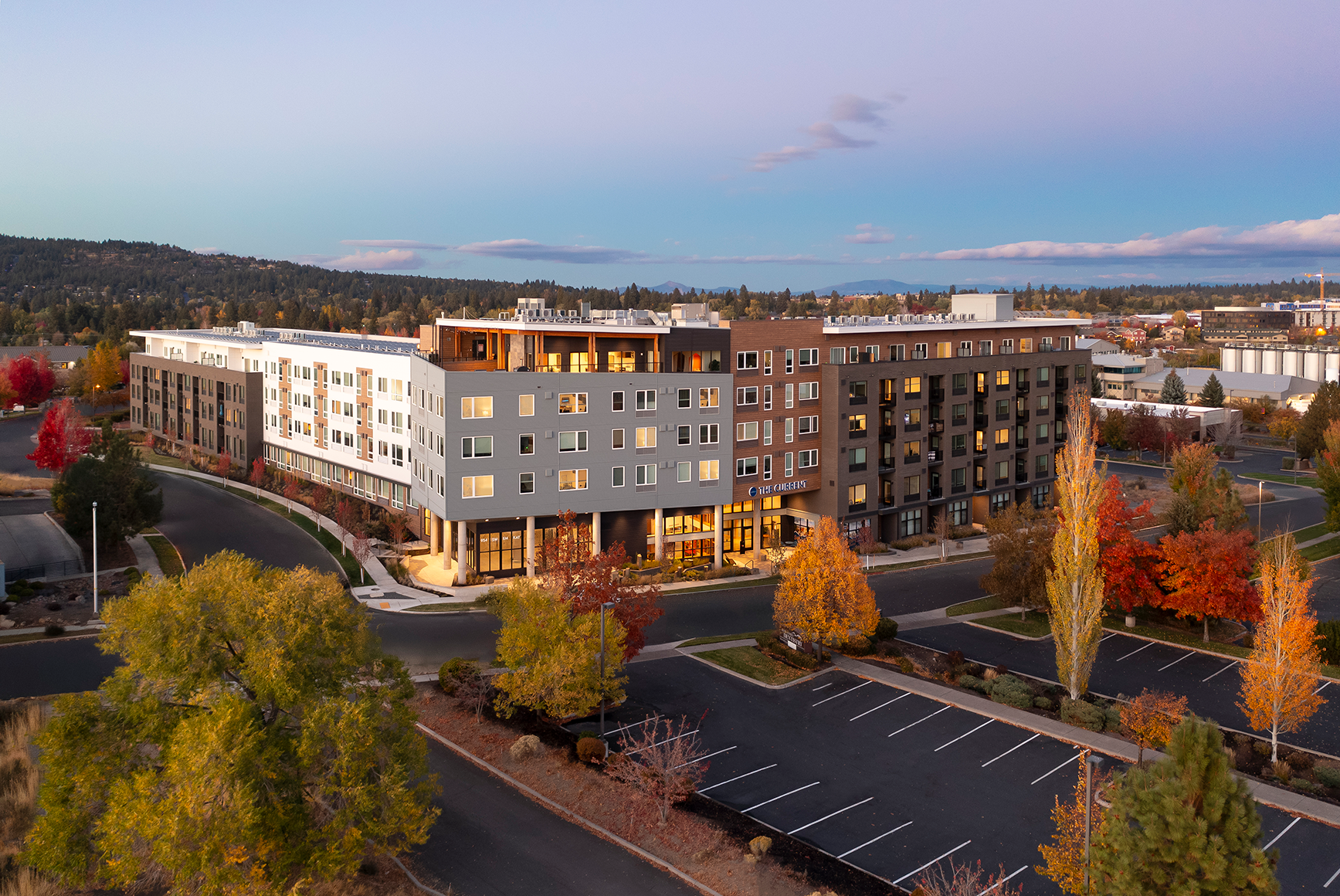 Modern apartment building called The Current surrounded by fall foliage and empty parking lots at dusk.