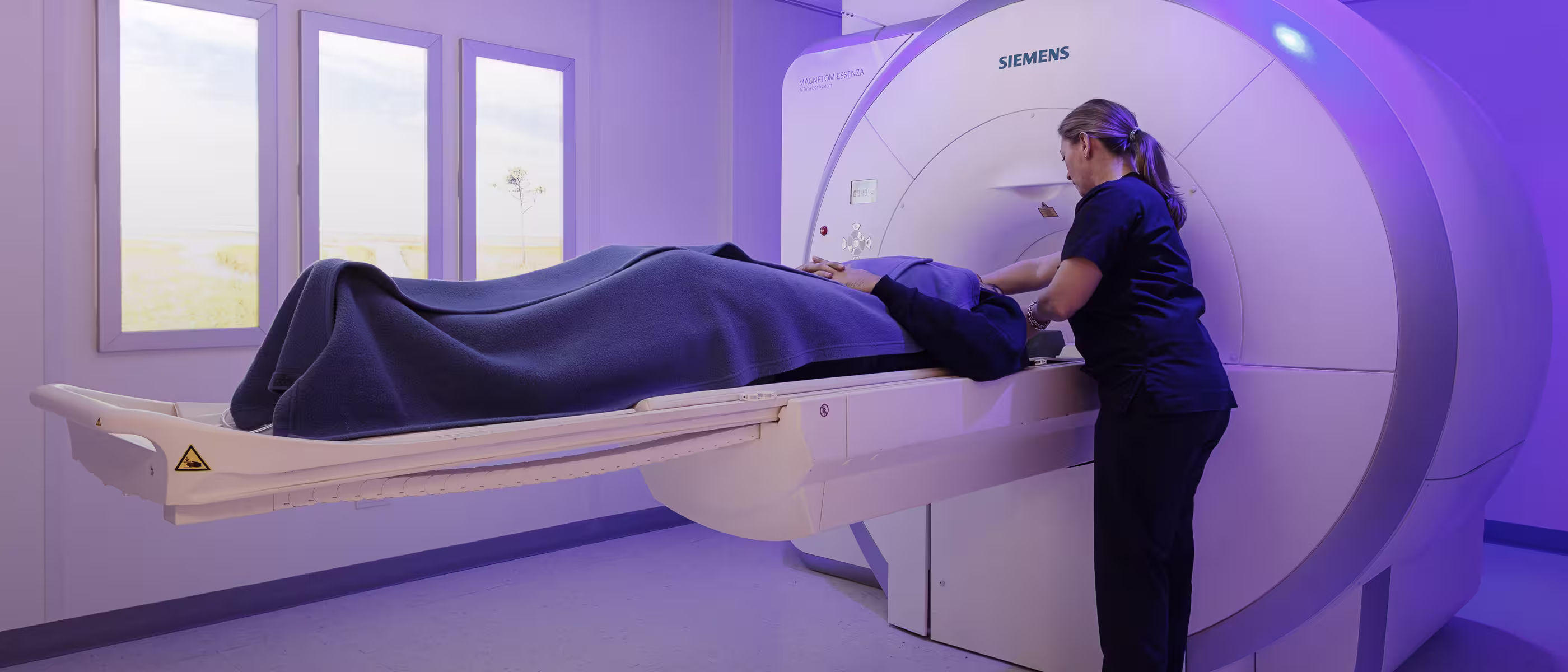 A medical professional adjusting a patient on a magnetic resonance imaging (MRI) machine in a clinical setting. The room has soft lighting and large windows.