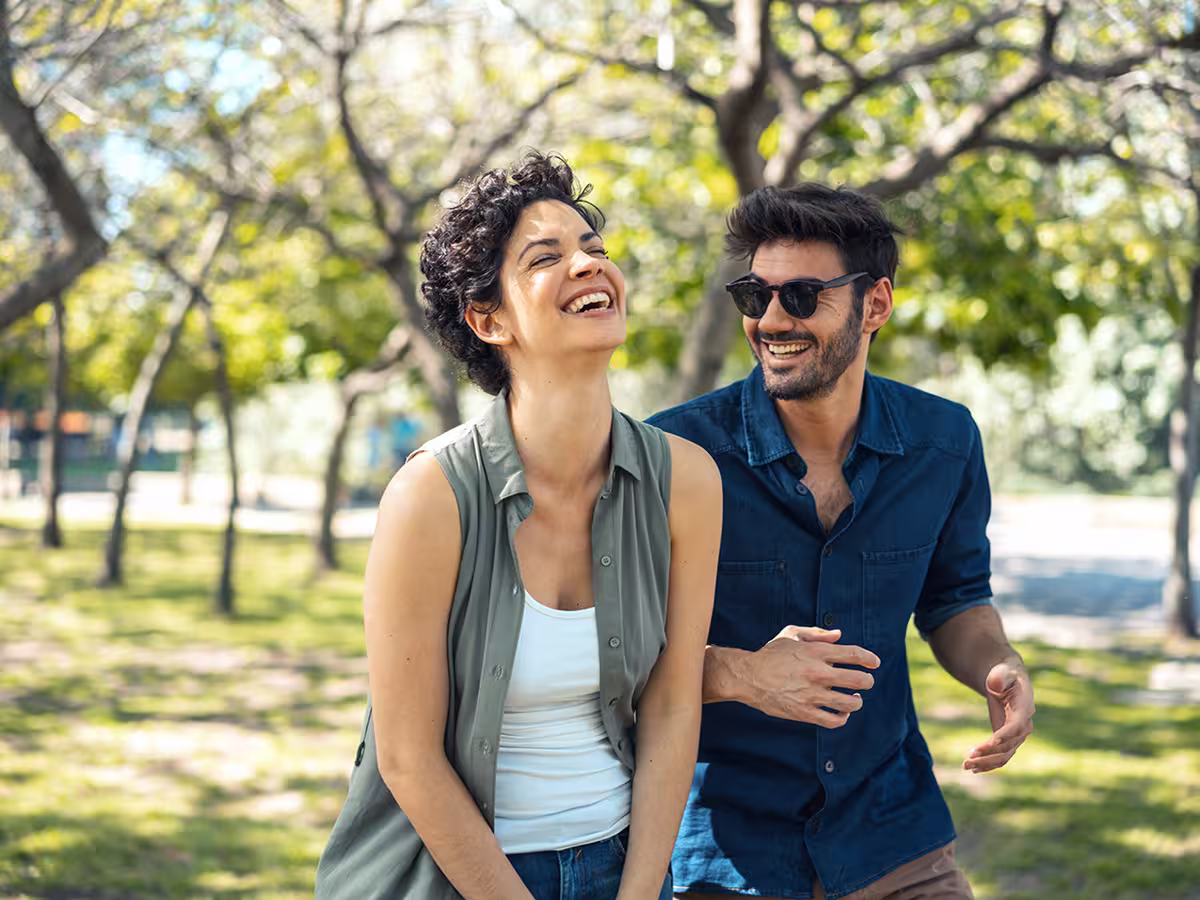 Pareja joven riendo juntos en un parque al aire libre con árboles y luz natural.