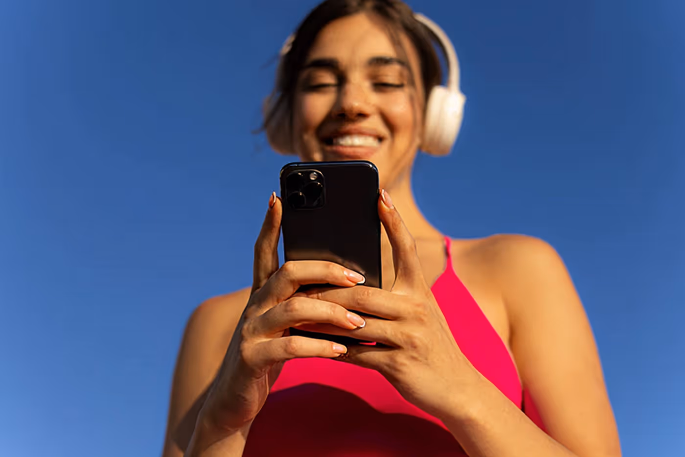 Mujer sonriente con auriculares blancos usando un celular negro bajo un cielo azul claro.