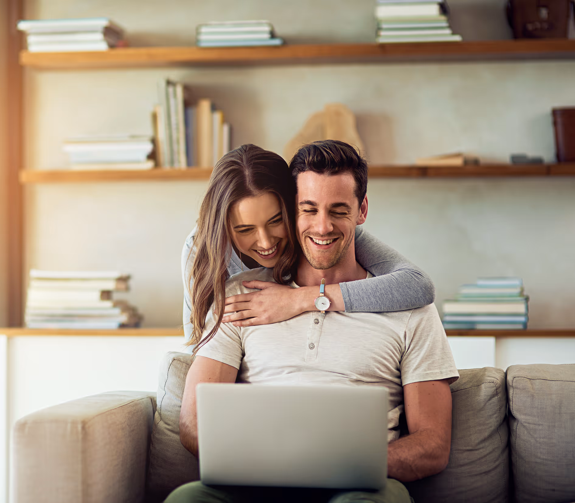 Pareja sonriente usando una computadora portátil en el sofá de una sala con estantes llenos de libros al fondo.