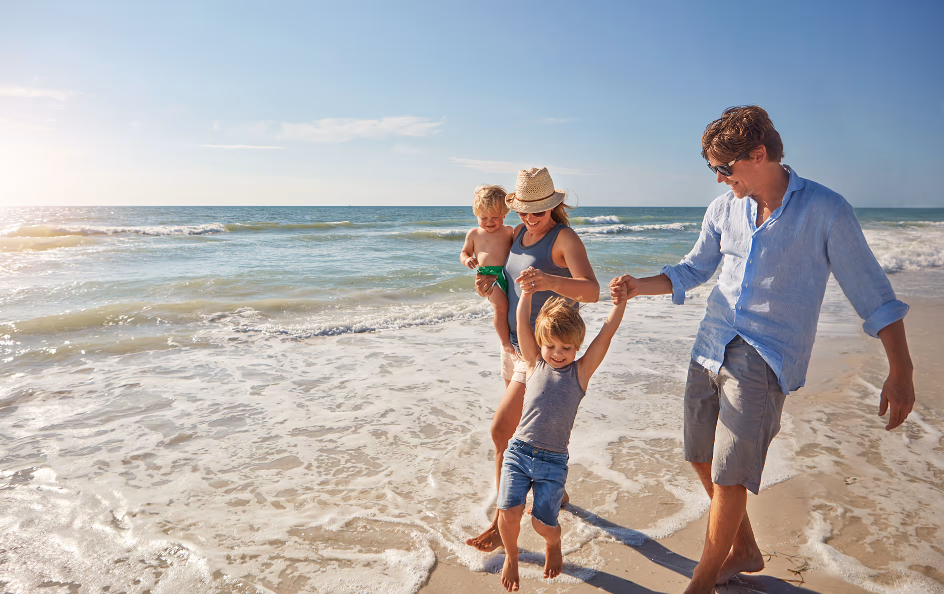 Familia con dos niños pequeños caminando y jugando en la orilla de una playa soleada con olas suaves.