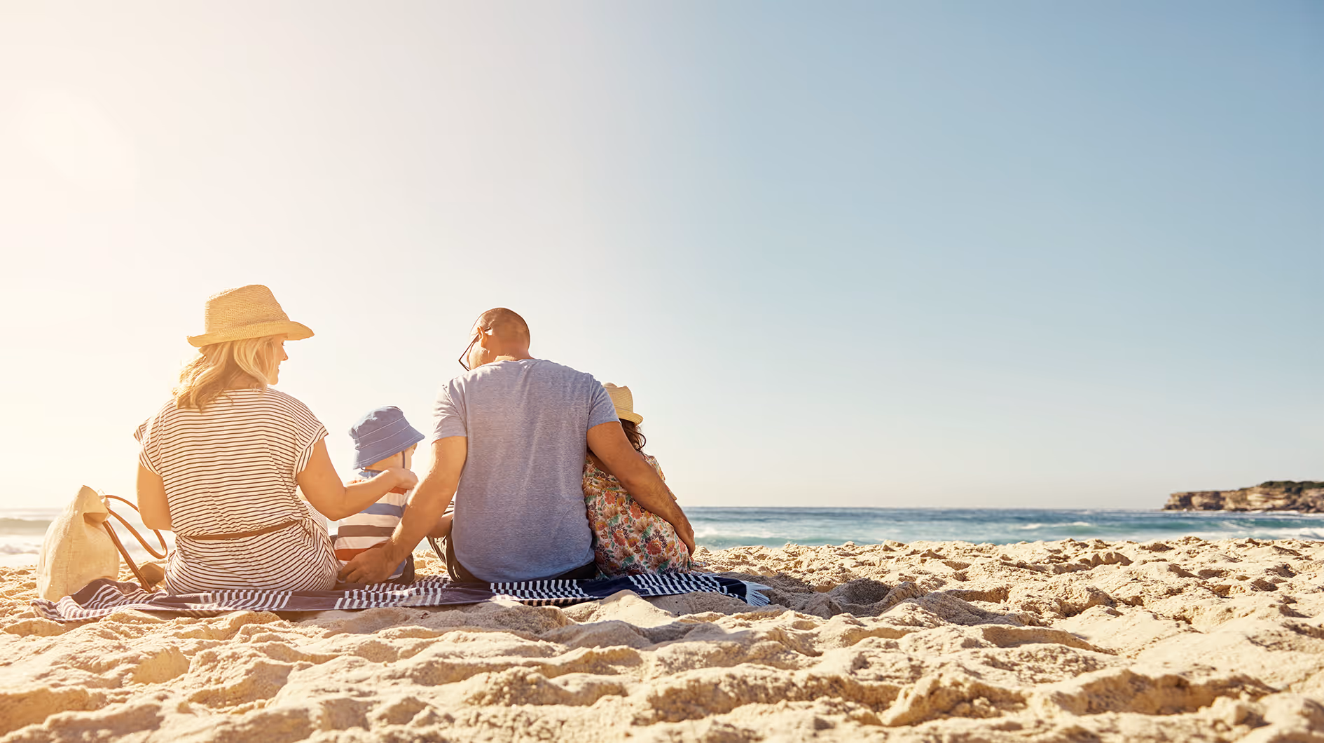 Familia sentada en una manta sobre la arena en la playa, mirando hacia el mar en un día soleado.