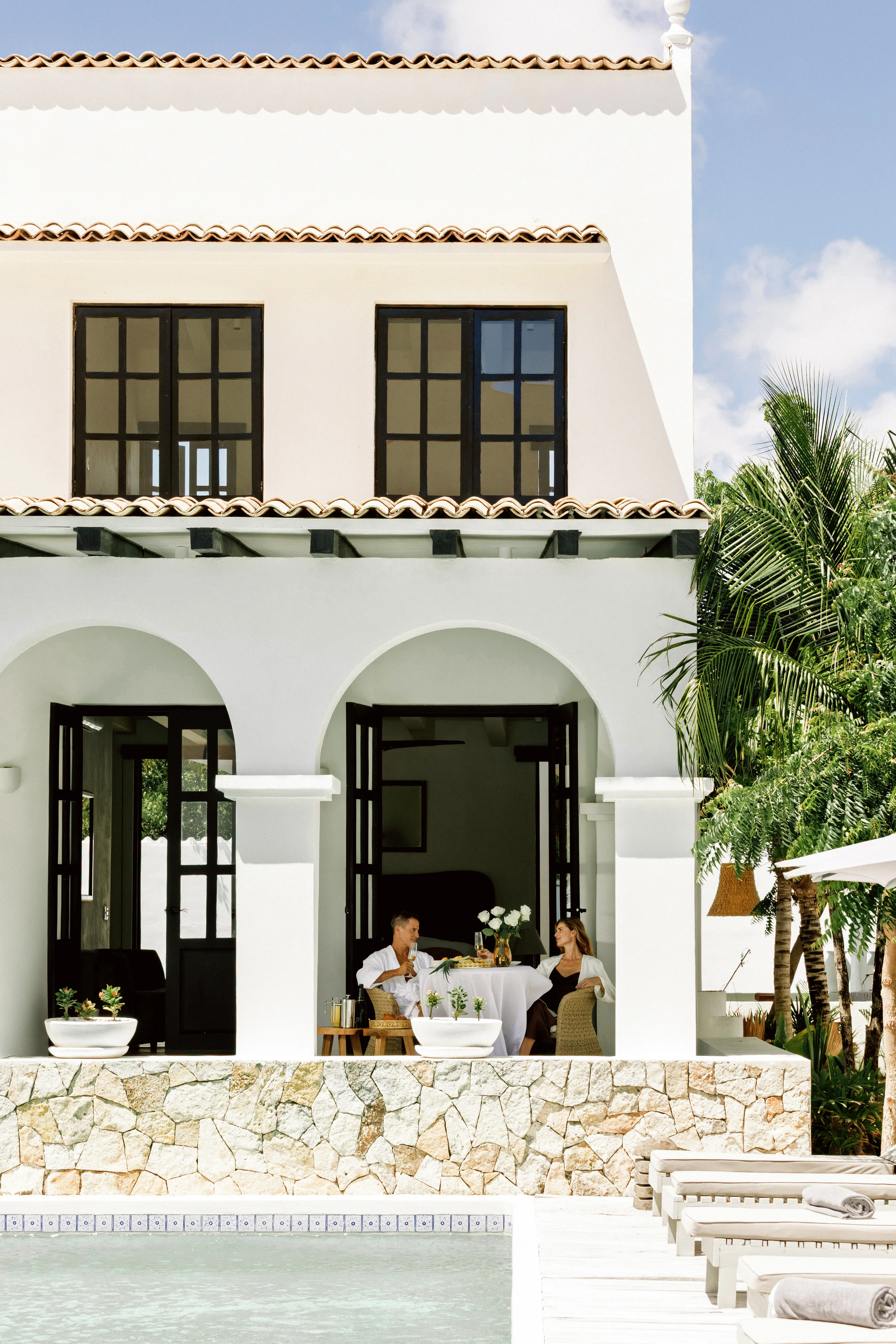 Couple sitting at a table with white tablecloth on a patio of a white villa with arches, next to a pool and surrounded by tropical plants.
