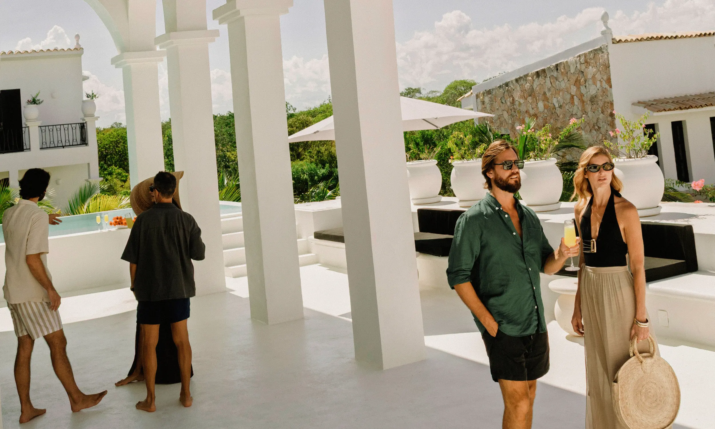 Four people in casual summer clothes socialize on a white terrace with large pillars in a sunny outdoor setting.
