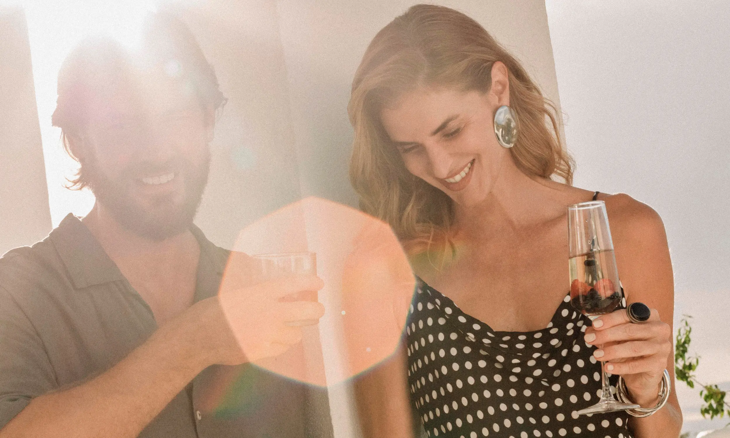 Smiling man and woman with drinks, sunlight creating lens flare effect.