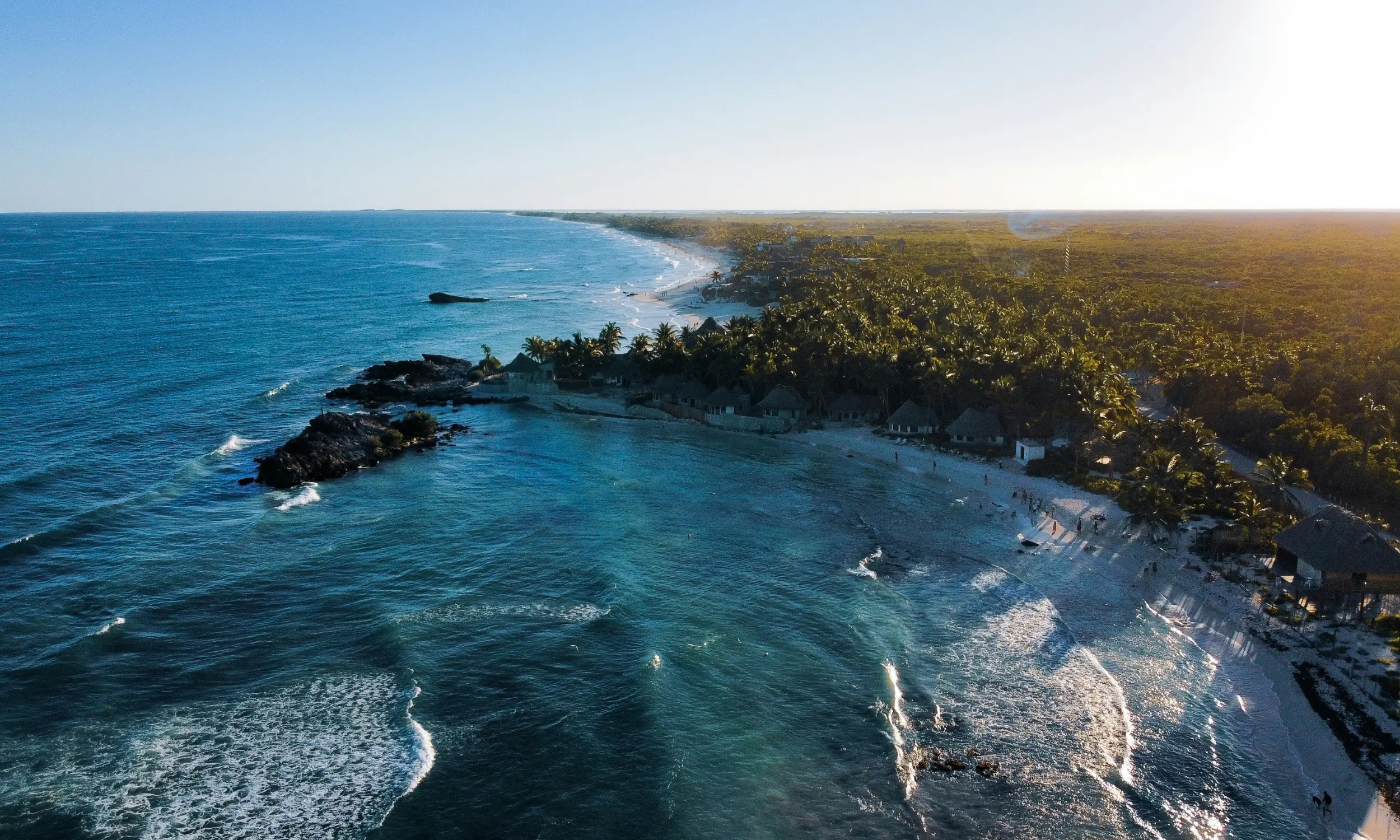 Aerial view of a tropical coastline with a sandy beach, palm trees, and small huts alongside the blue ocean under clear sky.