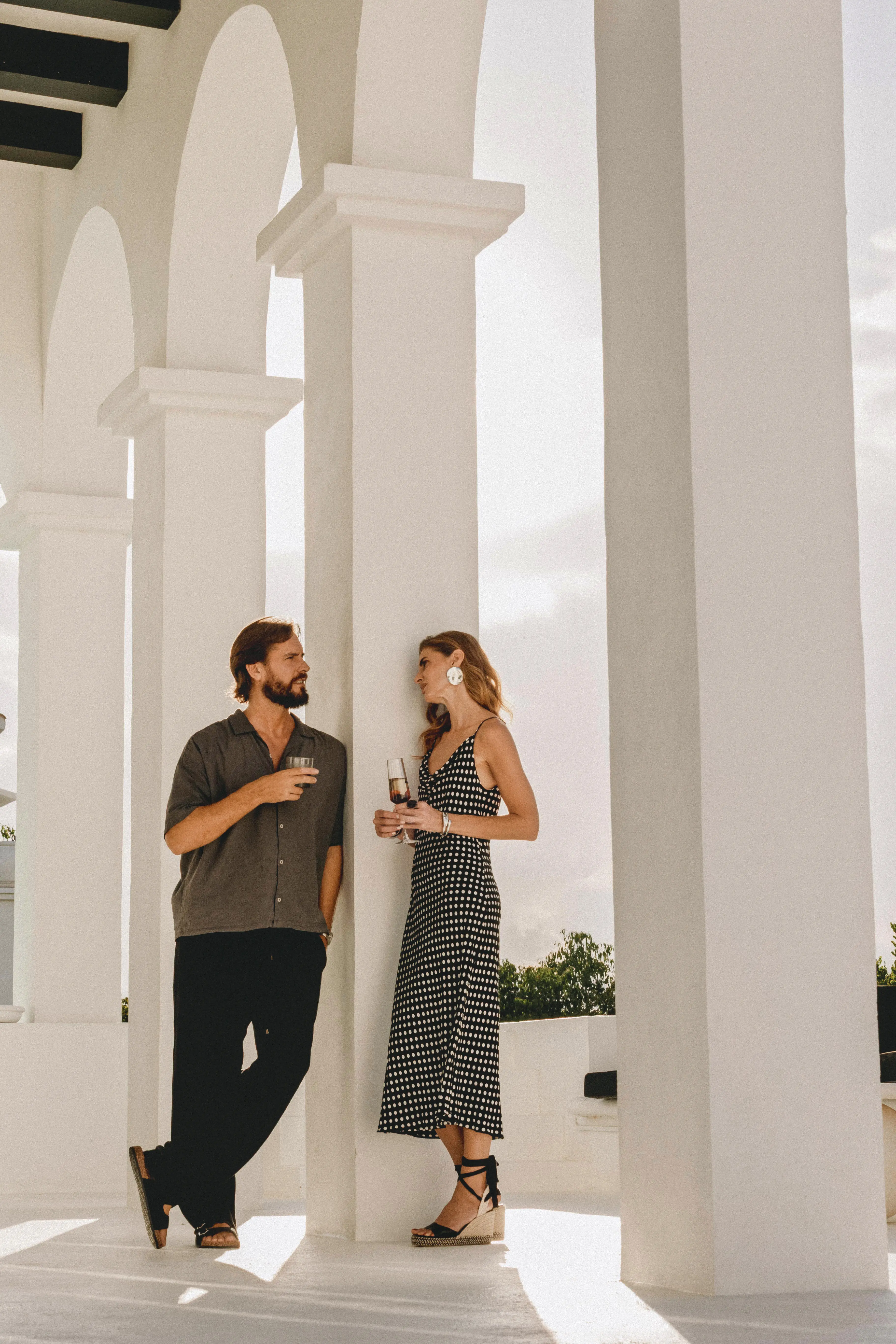 Man and woman standing near white columns, drinking wine and conversing outdoors in daylight.
