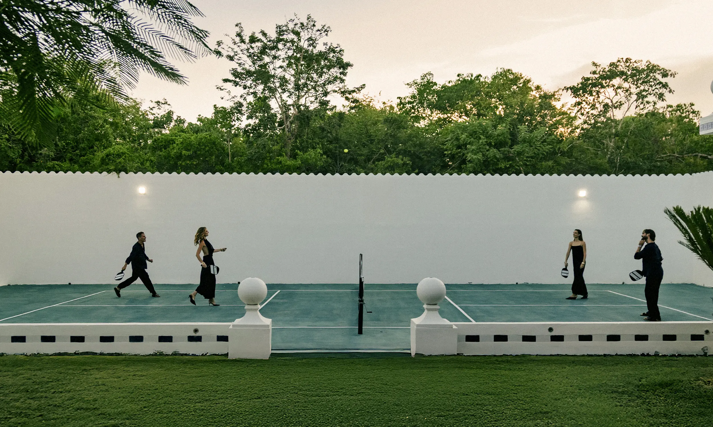 Four people playing pickleball on an outdoor court with a white wall and green trees in the background.