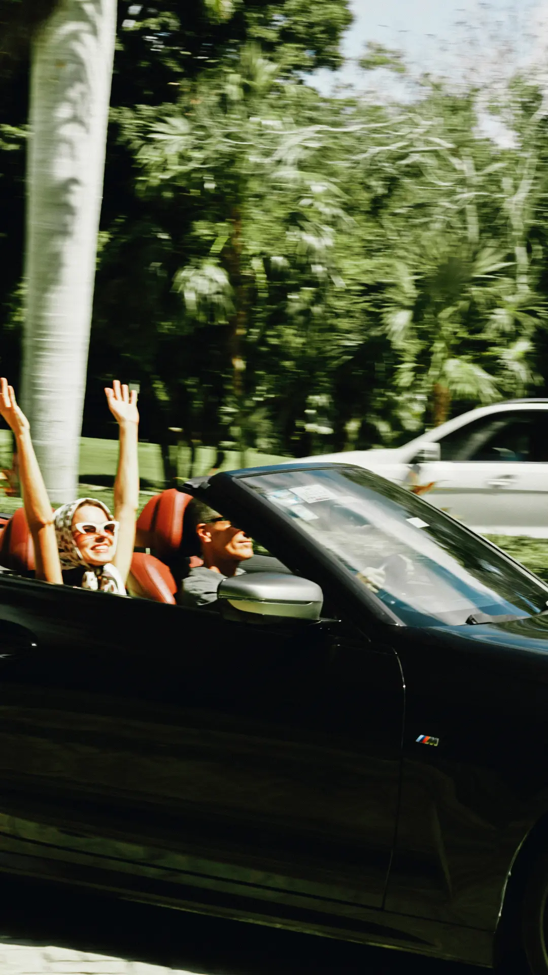 Woman with scarf and sunglasses joyfully raising arms in the passenger seat of a black convertible car driven by a man on a sunny day.