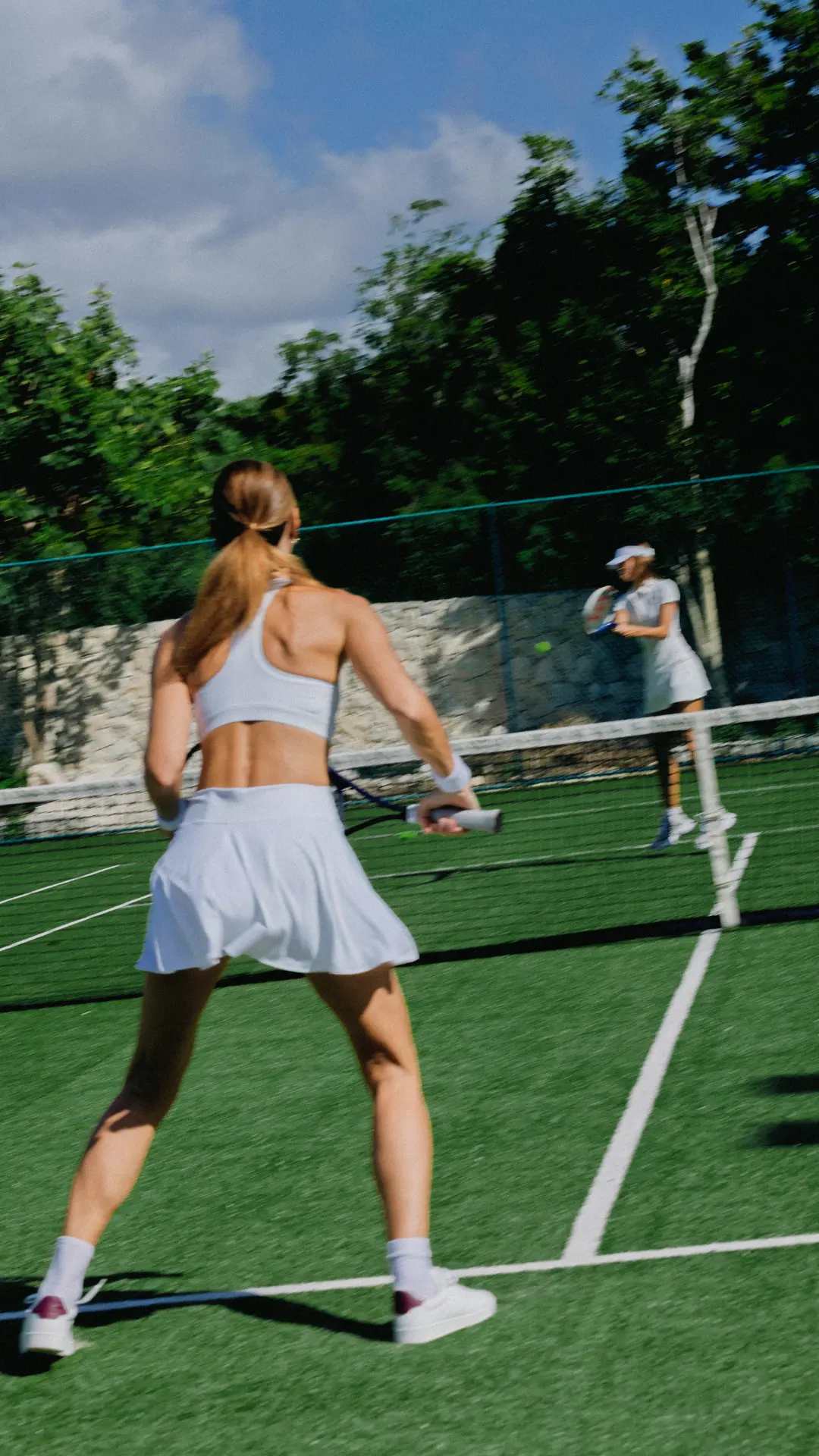 Two women playing tennis on an outdoor court surrounded by trees under a blue sky.